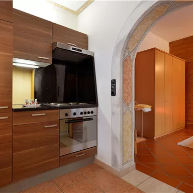 A modern kitchen with wooden cabinets and a stove. In the background, an adjacent room with a cozy bed is visible.