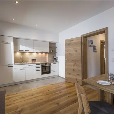 A modern kitchen with white cabinets and wooden shelves. The dining area is cozy with a wooden table and dishes.