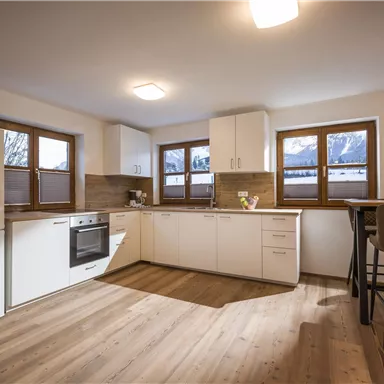 Eine moderne Küche mit weißen Schränken und großen Fenstern. Der Boden ist aus Holz und der Raum wirkt hell und einladend.