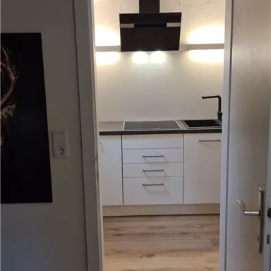 A modern kitchen with white furniture and a cooktop. The room is well-lit and has a wooden floor.