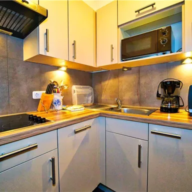 Eine moderne Küche mit hellen Schränken und einem Kochfeld. Sie ist ausgestattet mit einem Mikrowellenofen, einem Kaffeeautomaten und einer Spüle.