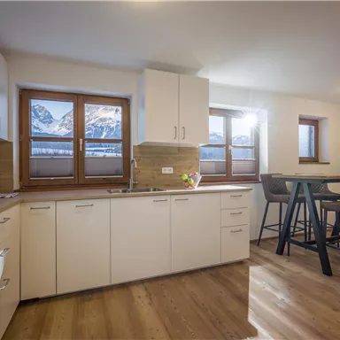 Eine moderne Küche mit Holzfußboden und großen Fenstern. Im Hintergrund sind schneebedeckte Berge zu sehen.