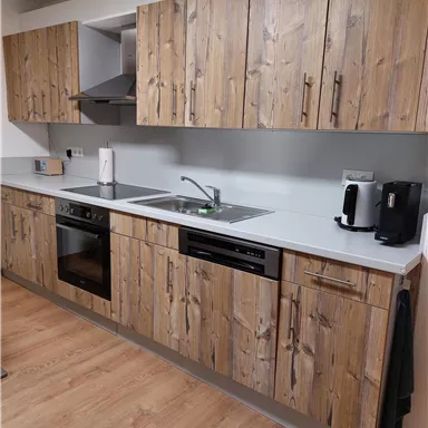 A modern kitchen with wooden fronts and a clear, minimalist design. The equipment includes a stove, a sink, and a coffee machine.