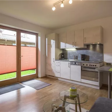 A modern kitchen with bright furniture and a large window front. The room has a cozy dining area and access to a garden.