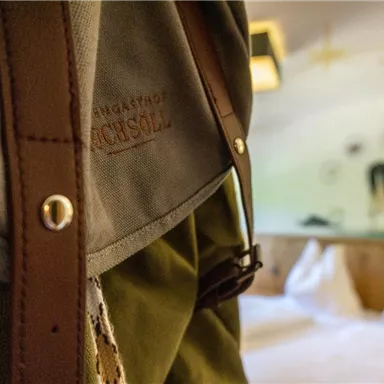 Ein naher Blick auf einen Rucksack mit Lederdetails. Im Hintergrund ist ein gemütliches Zimmer mit weißen Betten und einer Wandmalerei zu sehen.