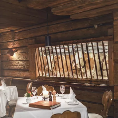 A rustic restaurant with wooden walls and elegant tables. Animals can be seen through the window grilles, offering a unique view.
