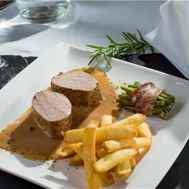 Ein köstliches Gericht mit zartem Fleisch, einer cremigen Soße und knusprigen Pommes frites. Dazu gibt es frische grüne Bohnen in Speck gewickelt und ein Glas Wein im Hintergrund.