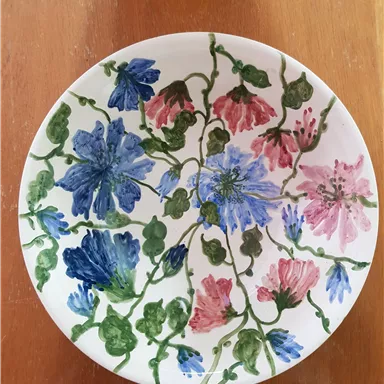 A decorative plate with colorful flower motifs in blue, pink, and green. The flowers are artfully entwined, giving the plate a fresh, nature-connected look.