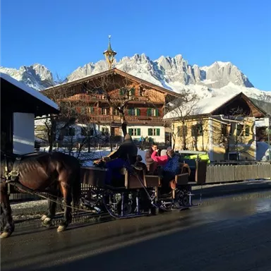 Eine malerische Winterszene mit einem traditionellen Haus und schneebedeckten Bergen im Hintergrund. Eine Pferdeschlittenfahrt verleiht der Landschaft einen charmanten, rustikalen Charakter.