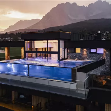 Ein modernes Haus mit einem Infinity-Pool und großen Fenstern. Im Hintergrund sind majestätische Berge und ein schöner Sonnenuntergang zu sehen.