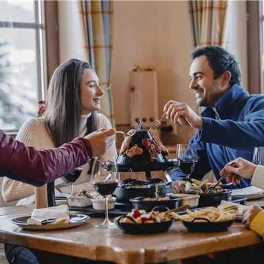 Eine Gruppe von vier Freunden sitzt an einem Tisch und genießt ein Essen miteinander. Die Atmosphäre ist gemütlich und einladend, mit Wein und verschiedenen Gerichten auf dem Tisch.