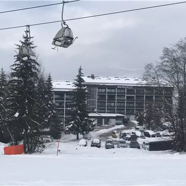 A hotel in a snow-covered landscape with fir trees. In the foreground, ski lifts and a parking lot can be seen.