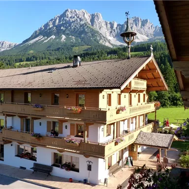 Ein traditionelles alpenländisches Holzhaus mit Balkonen, umgeben von grünen Wiesen und majestätischen Bergen. Der Himmel ist klar und die Umgebung lädt zur Entspannung ein.