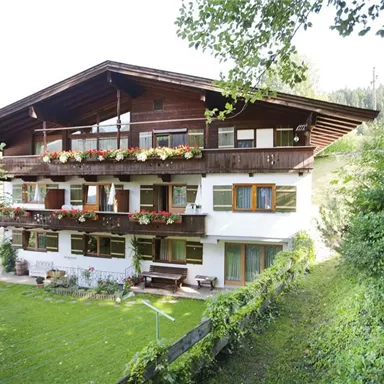 A charming alpine-style house with several balconies and colorful flowers. Surrounded by lush greenery and trees, it conveys an inviting atmosphere.