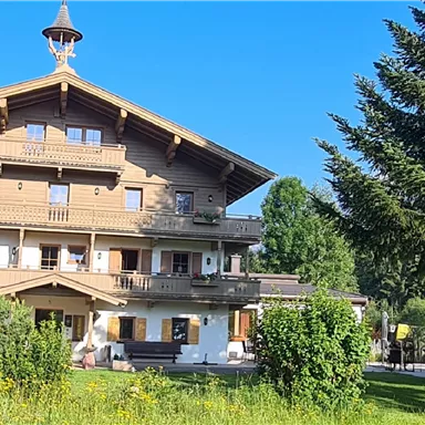 Ein charmantes Holzgebäude im alpinen Stil unter einem klaren blauen Himmel. Umgeben von Grünflächen und Bäumen.
