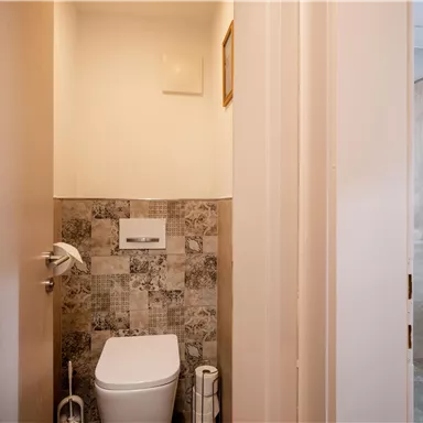 A small bathroom with a toilet and a gray tiled wall. Light comes in through the window into the upper area of the room.
