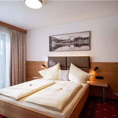 A cozy bedroom with a large bed, decorated pillows, and wooden furniture. The large windows let in plenty of daylight.