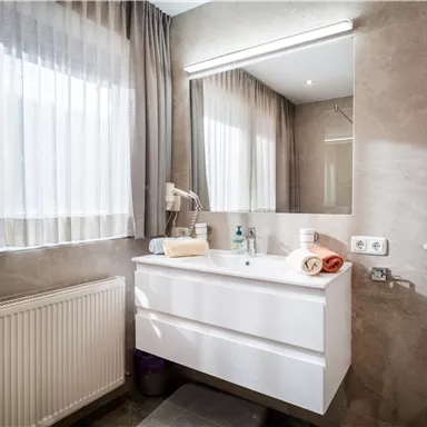 A modern bathroom with a simple white washbasin and a large mirror. The light filters through the curtains, creating a bright, inviting atmosphere.