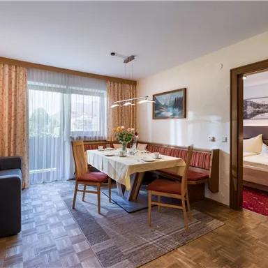 Ein gemütlicher Wohnbereich mit einem Esstisch und Stühlen sowie einem Fernseher. Durch die großen Fenster fällt viel Licht in den Raum.