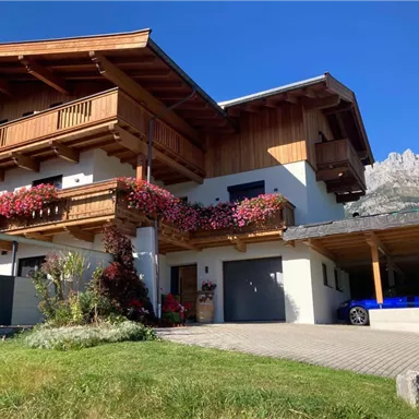 Ein schönes Holzhaus mit Blumenkästen und einem geräumigen Eingang. Im Hintergrund sind Berge und ein blauer Himmel zu sehen.