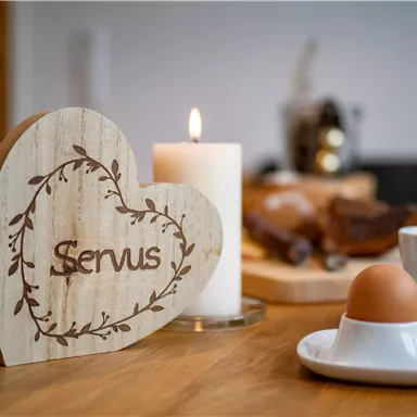 A heart-shaped wooden sign with the inscription "Servus" is standing on a table. In the background, candlelight, a boiled egg, and various breakfast foods are visible.