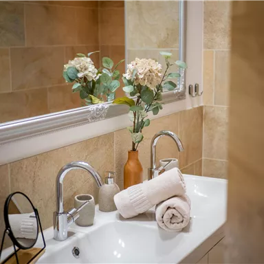 A modern bathroom with a white sink and elegant fixtures. Fresh flowers in a vase and some towels are on the shelf.