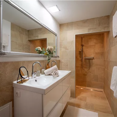 A modern bathroom with a bright sink and a large mirror. The walls are designed with warm beige tiles and there is a shower in the background.