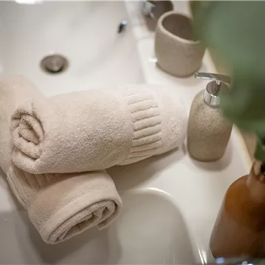 Soft, beige towels are lying on a sink. In the background, care products and a plant can be seen.