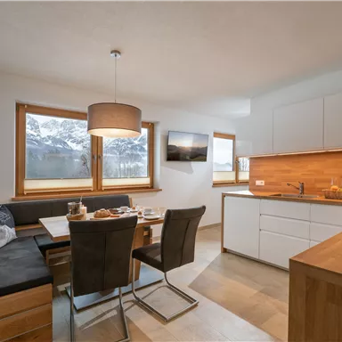 A modern kitchen with a dining area and large windows. The view of the mountains is visible and creates a bright, inviting atmosphere.