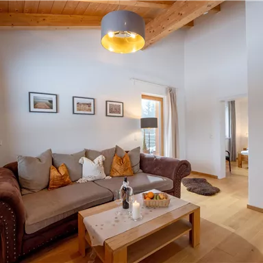A cozy living room with a beige sofa and decorative cushions. A coffee table with fruits and a bottle is at the center of the room.