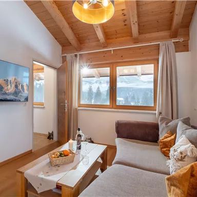 A cozy living room with wooden beam ceiling and large windows. The atmosphere is inviting, with a sofa and a table holding fresh fruit.