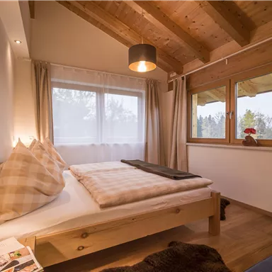 A cozy bedroom with a wooden ceiling and large windows. The room is simple but inviting, with a bed and decorative pillows.