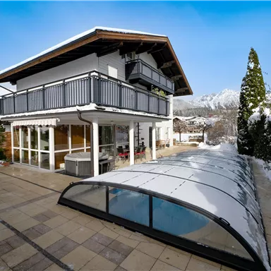 A modern house with a balcony in a snowy landscape. The outdoor area features a covered pool and surrounding mountains in the background.