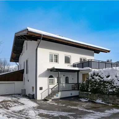 A modern house in the snow with a large, covered balcony. The surroundings are surrounded by snow-covered mountains and the sky is clear and blue.