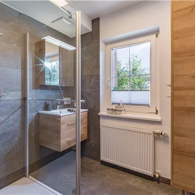 A modern bathroom with a glass shower and wooden details. The window lets in plenty of light and offers a view outside.