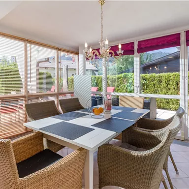 A bright conservatory with a dining area. The table is set with a decorative tablecloth and surrounded by cozy chairs.