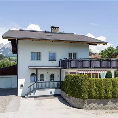 A beautiful, modern house with a balcony and a green garden. In the background, mountains and a blue sky can be seen.