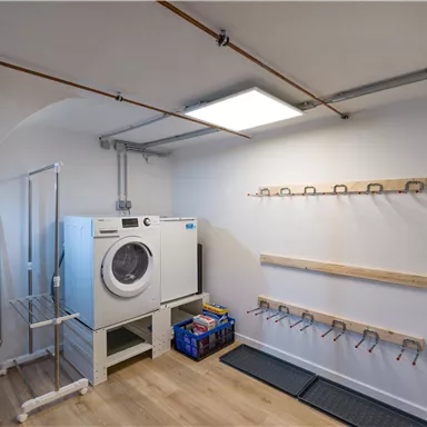 A bright laundry room with a washing machine and a drying rack. Coat hooks are attached to the wall.