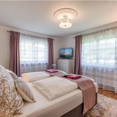 A bright and modern bedroom with two beds and soft curtains. A TV and an elegant carpet complement the cozy atmosphere.
