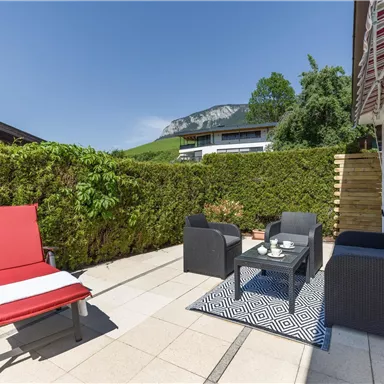 A cozy terrace with modern furniture and a view of the surrounding nature. The area is surrounded by green bushes and mountains.