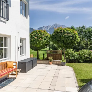 A bright terrace with a wooden chair and a seating area. In the background, green trees and majestic mountains can be seen.