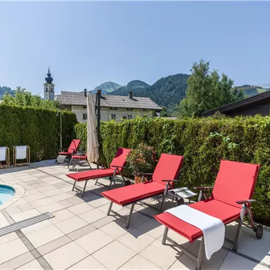 A sunny pool area with red loungers and a green hedge. In the background, mountains and a church tower can be seen.