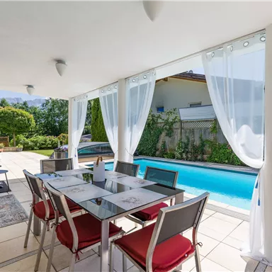 An elegant dining area with a glass table and chairs next to a pool. The light curtains create an inviting atmosphere outdoors.