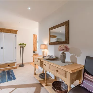 An inviting entryway with a wooden table, a lamp, and a small vase. A mirror hangs on the wall, and a comfortable chair stands next to it.