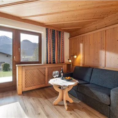 Ein gemütliches Wohnzimmer mit Holzwänden und einem Sofa. Große Fenster bieten einen Blick auf die Berge und das Freie.