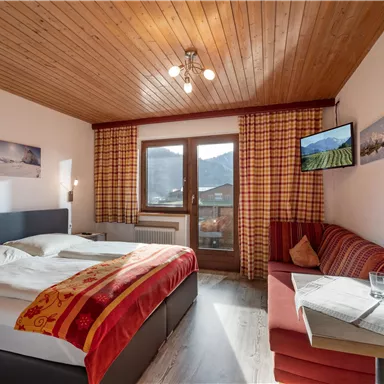 Ein gemütliches Hotelzimmer mit zwei Betten und einer roten Couch. Große Fenster bieten einen Blick auf die Berge und der Raum ist hell und einladend.