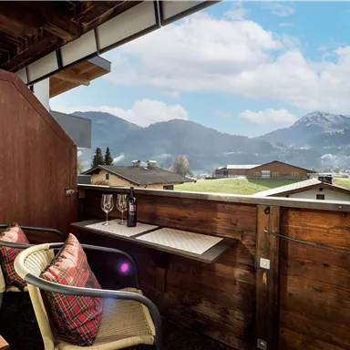 Ein gemütlicher Balkon mit zwei Stühlen und einem Tisch, der Blick auf grüne Hügel und Berge bietet. Die Atmosphäre ist einladend und ideal zum Entspannen.