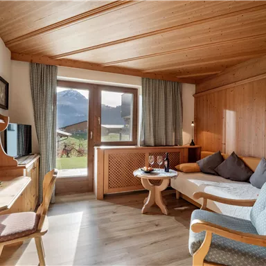 Ein gemütliches Zimmer mit Holzwänden und einem Fenster, das einen Blick auf die Berge bietet. Im Raum stehen ein Tisch, eine Couch und ein Schreibbereich.