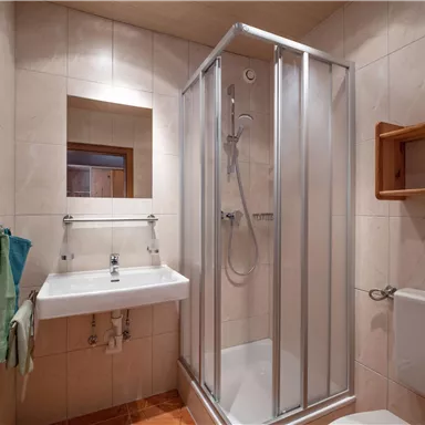 Ein modernes Badezimmer mit einer Duschkabine und einem Waschbecken. Die Wände sind hell gefliest und es gibt Handtücher in sanften Farben.