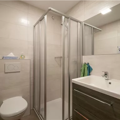 Ein modernes Badezimmer mit einer Dusche und einem Waschbecken. Die Wände sind hell gefliest und es gibt praktische Ablageflächen.